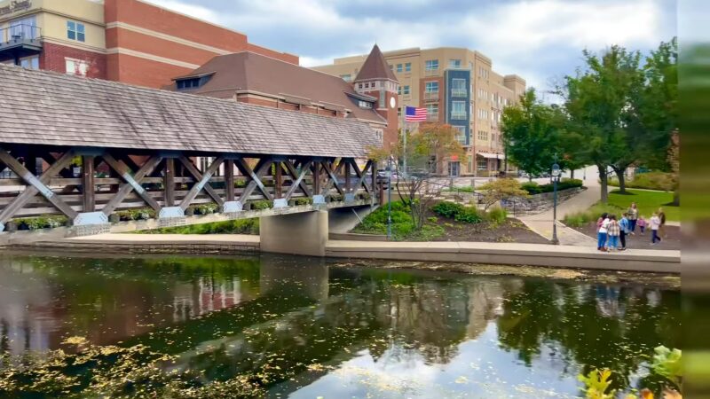 View of Naperville Featuring a Reflective River, a Covered Bridge with White Benches, Surrounded by Multi-Story Buildings and A Vibrant Green Landscape