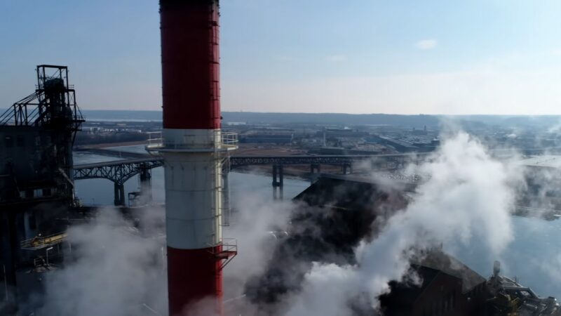 Aerial View of An Industrial Area in Peoria with A Prominent Red and White Chimney Emitting Steam, Overlooking a River and Bridges